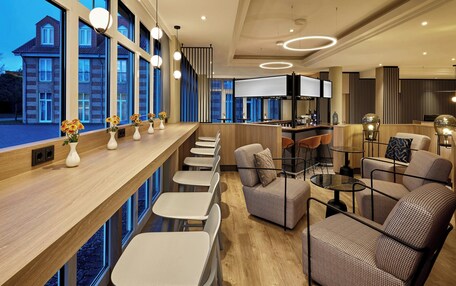 A modern hotel lobby with a long reception desk, seating area, and large windows offering a view of the building outside.