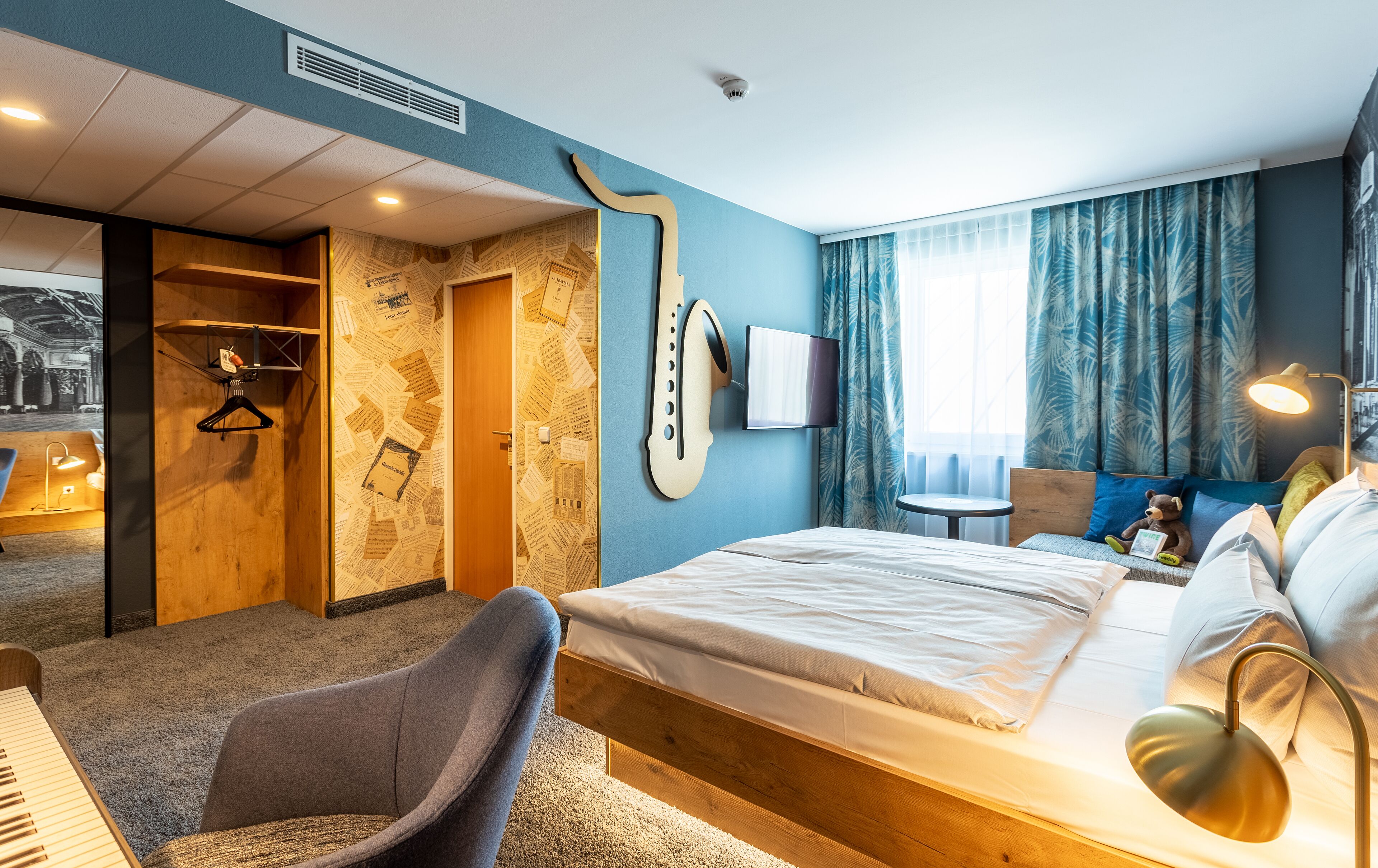 Ein Hotelzimmer mit Bett, Schreibtisch mit Tastatur, Stuhl, Kleiderhaken und einem an der Wand befestigten Fernseher, sowie einem Fenster mit Vorhängen.