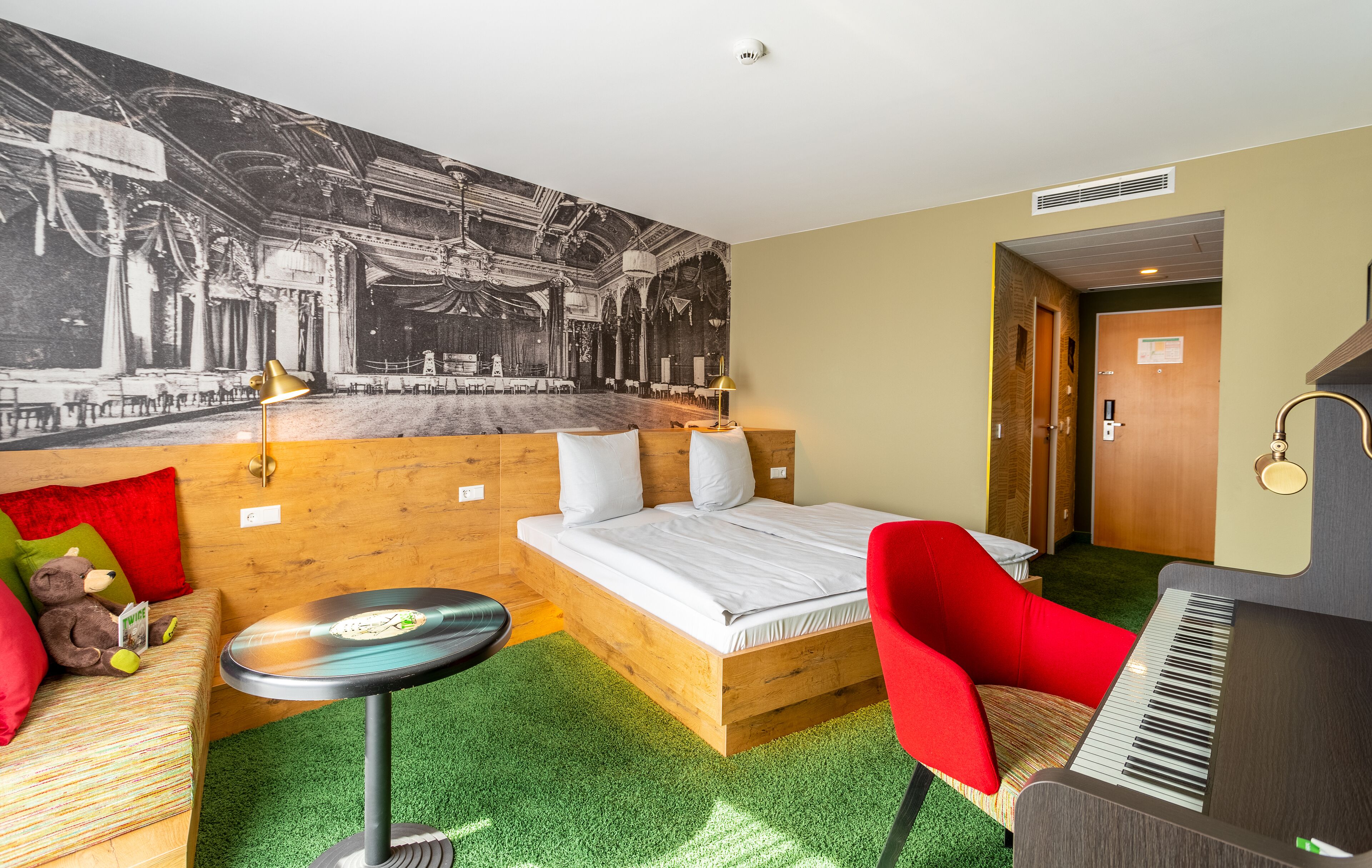 A hotel room with a bed, a red chair, a small table, a keyboard, and a mural on the wall.