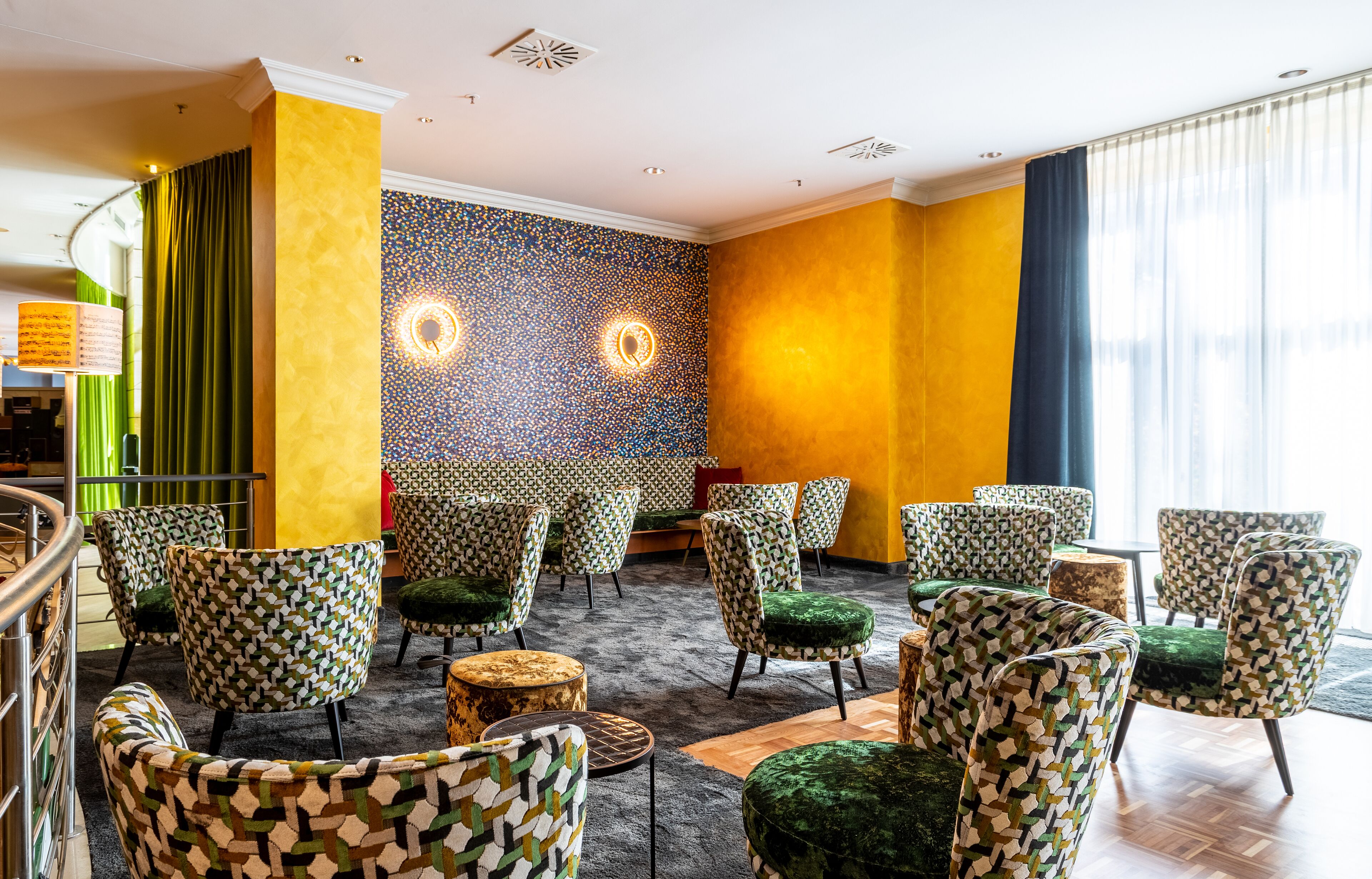 A hotel lobby with patterned chairs, a carpeted floor, a wall with a circular light fixture, and a staircase.