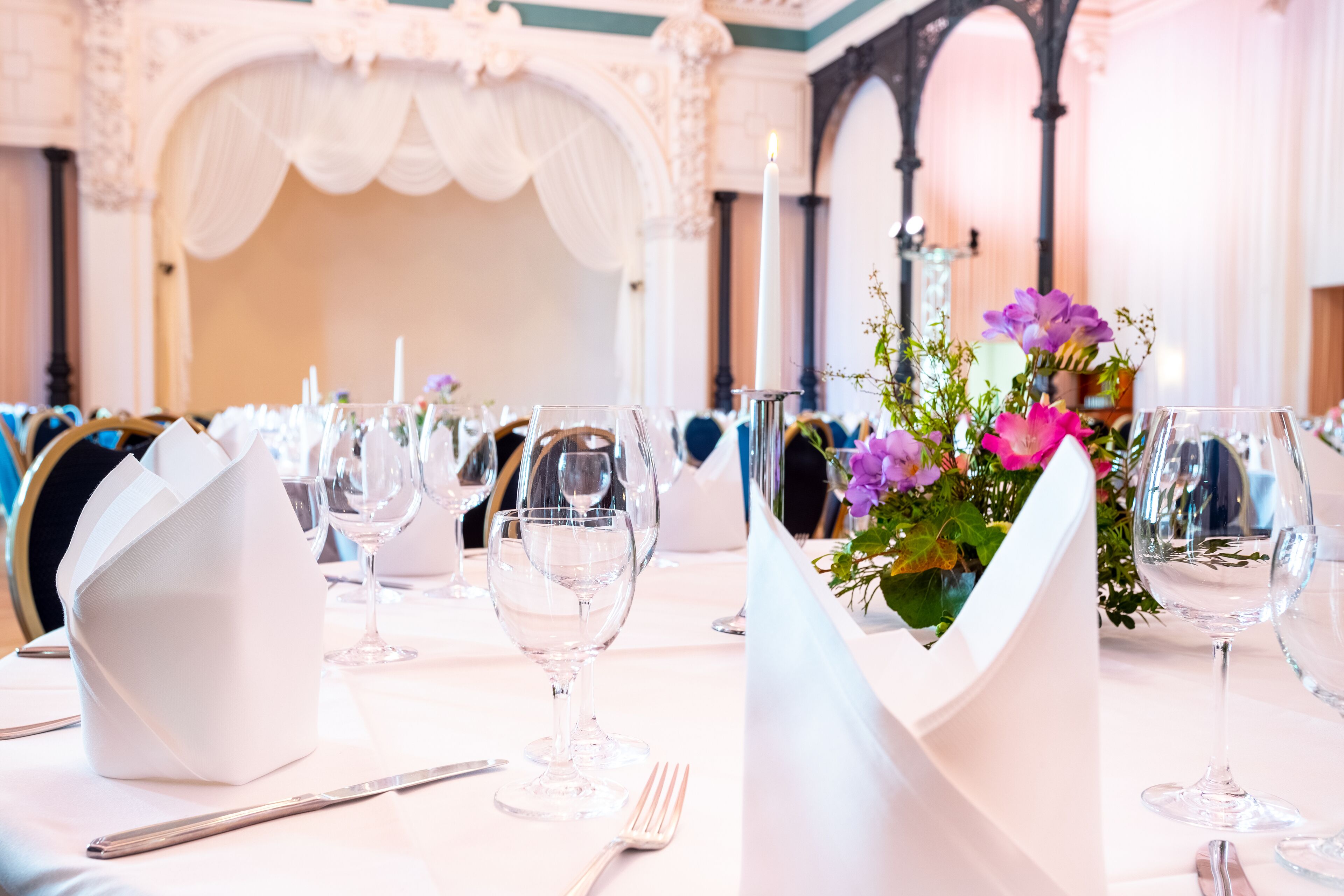 A banquet table set with white tablecloths, glassware, and a centerpiece with purple flowers.