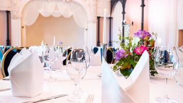 Una mesa de banquete dispuesta con manteles blancos, cristalería y un centro de mesa con flores moradas.