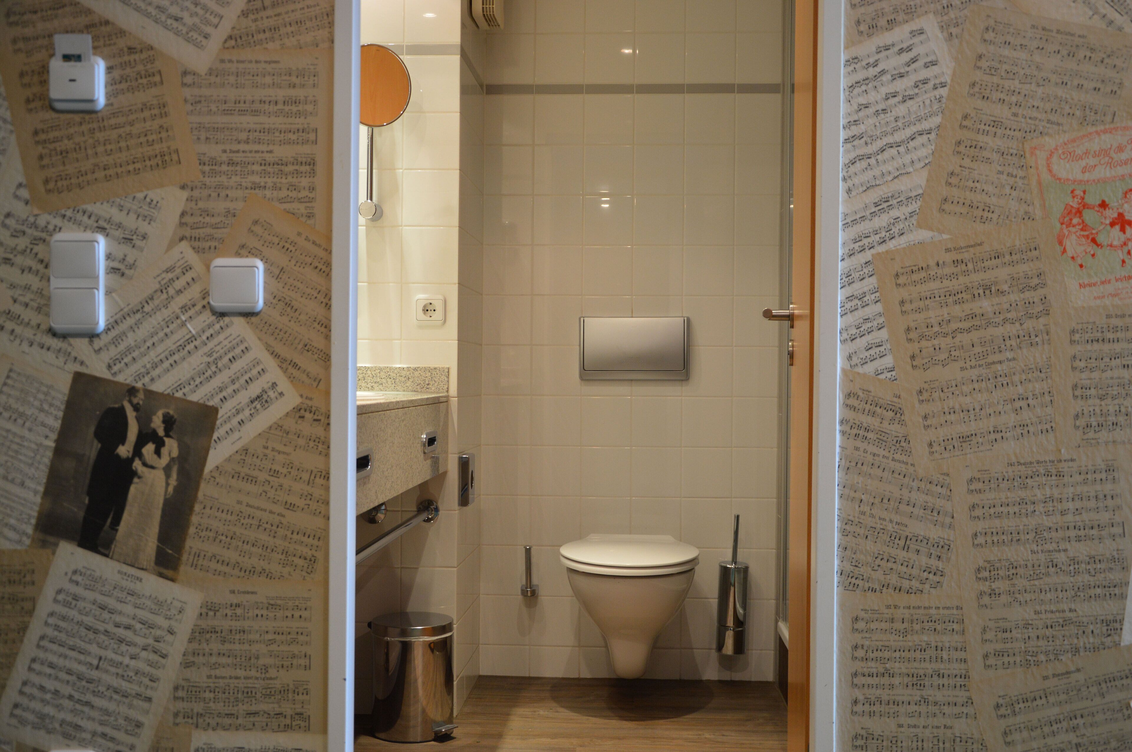 Ein Badezimmer mit Toilette, Waschbecken und Mülleimer. An den Wänden befinden sich Papiere und Fotos.