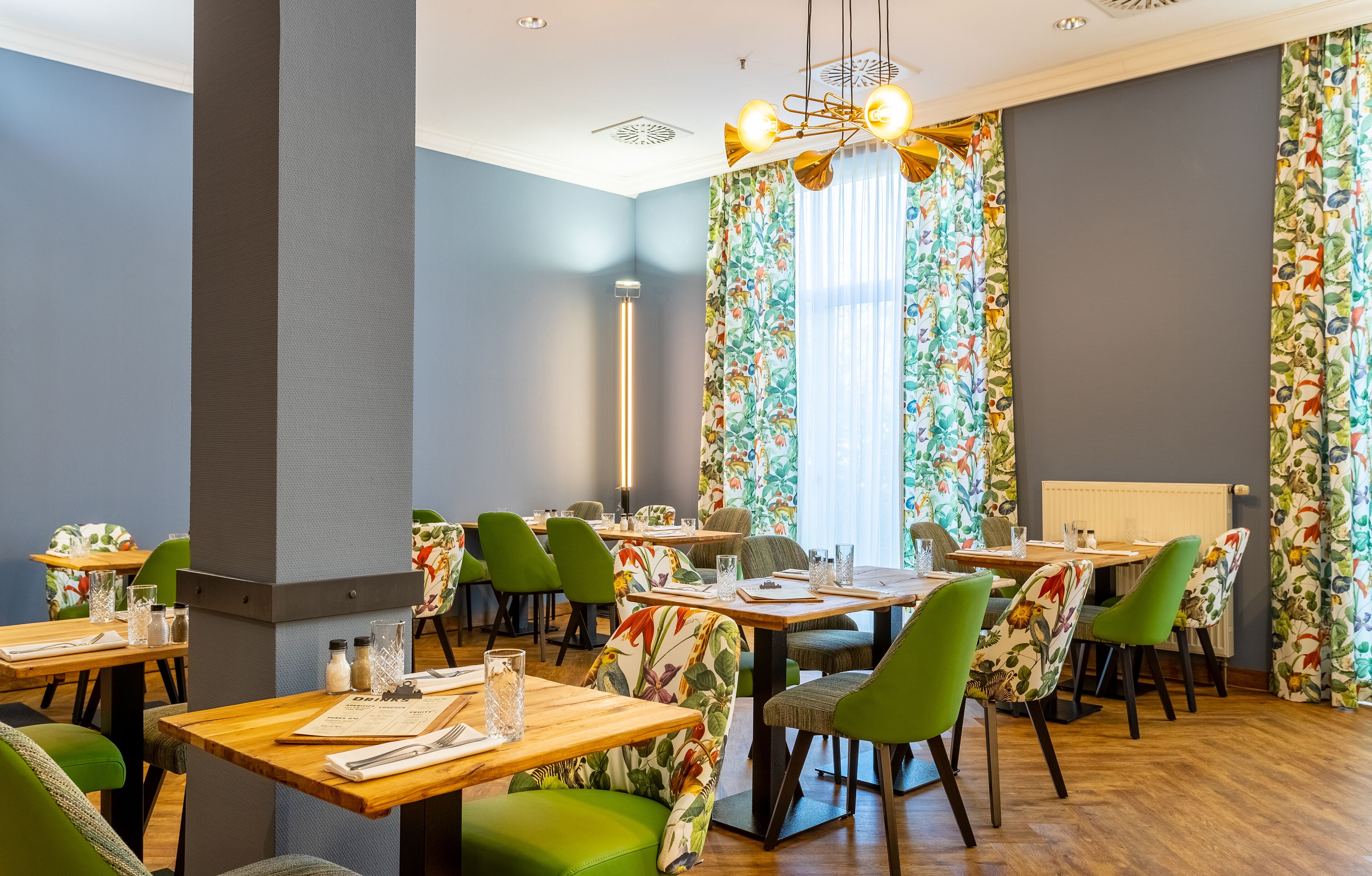Un restaurante con mesas de madera y sillas verdes, amplias ventanas con cortinas florales y una lámpara de araña moderna.