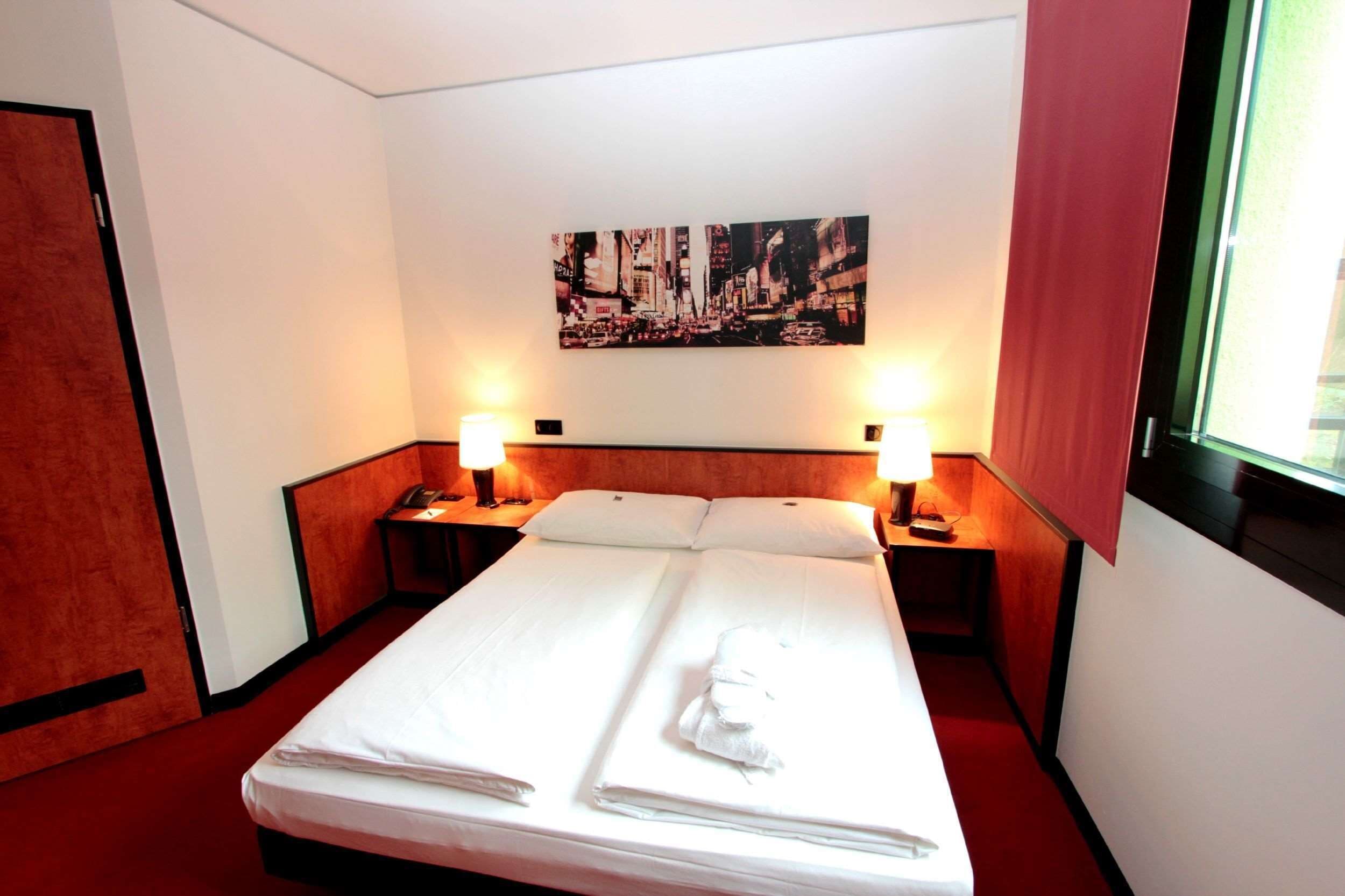 Ein Hotelzimmer mit einem Bett, zwei Nachttischen mit Lampen, einem Bild an der Wand und einem Fenster mit Vorhängen.
