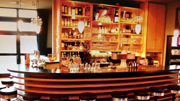 A bar area with a curved counter, multiple shelves stocked with bottles, and bar stools. - ARA-Hotel Comfort (Ingolstadt)