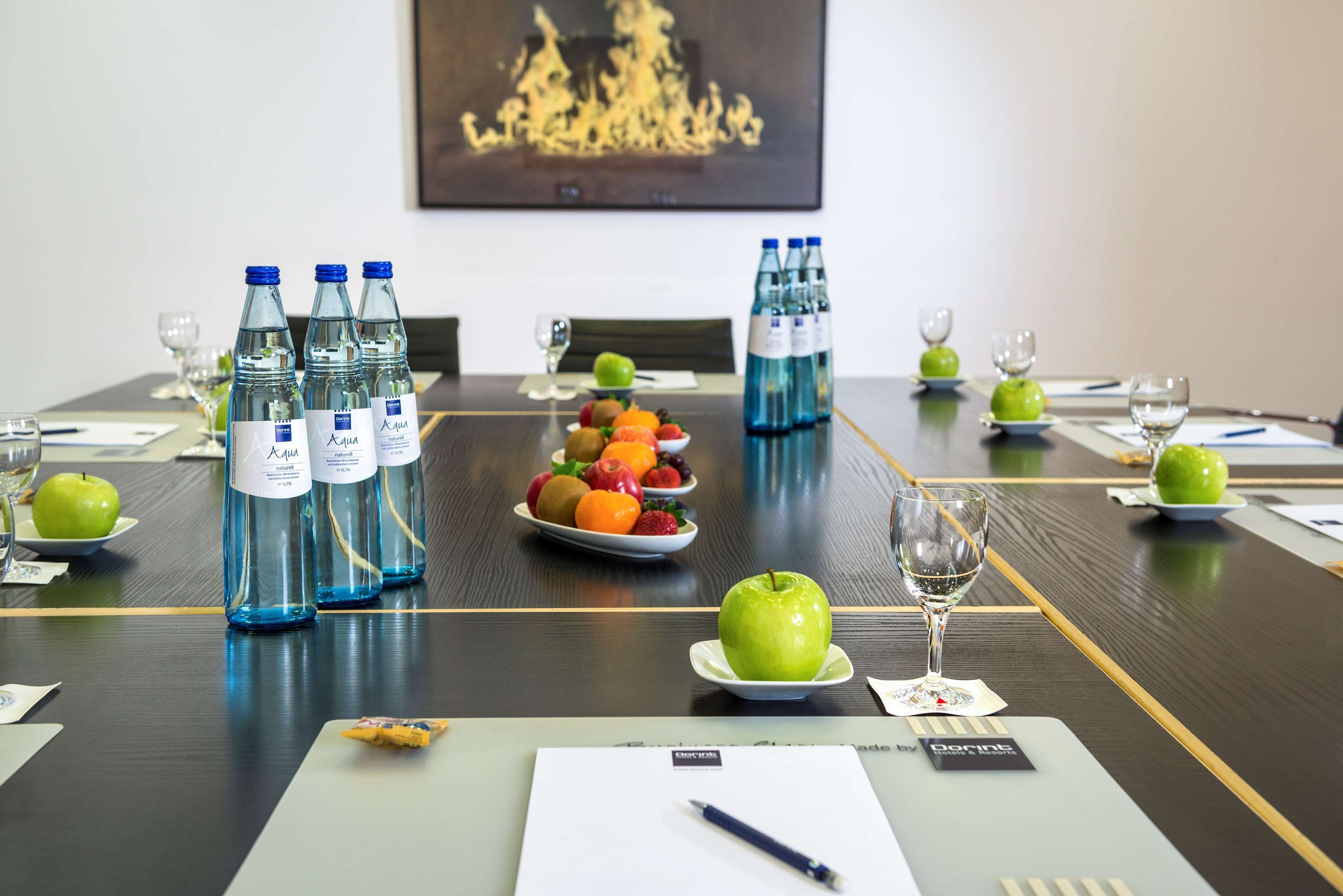 Mesa de conferencias preparada con botellas de agua, fruta y copas de vino, lo que sugiere una preparación para una reunión o evento.