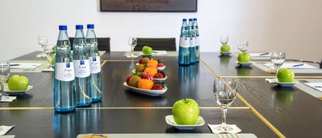 Mesa de conferencias preparada con botellas de agua, fruta y copas de vino, lo que sugiere una preparación para una reunión o evento.