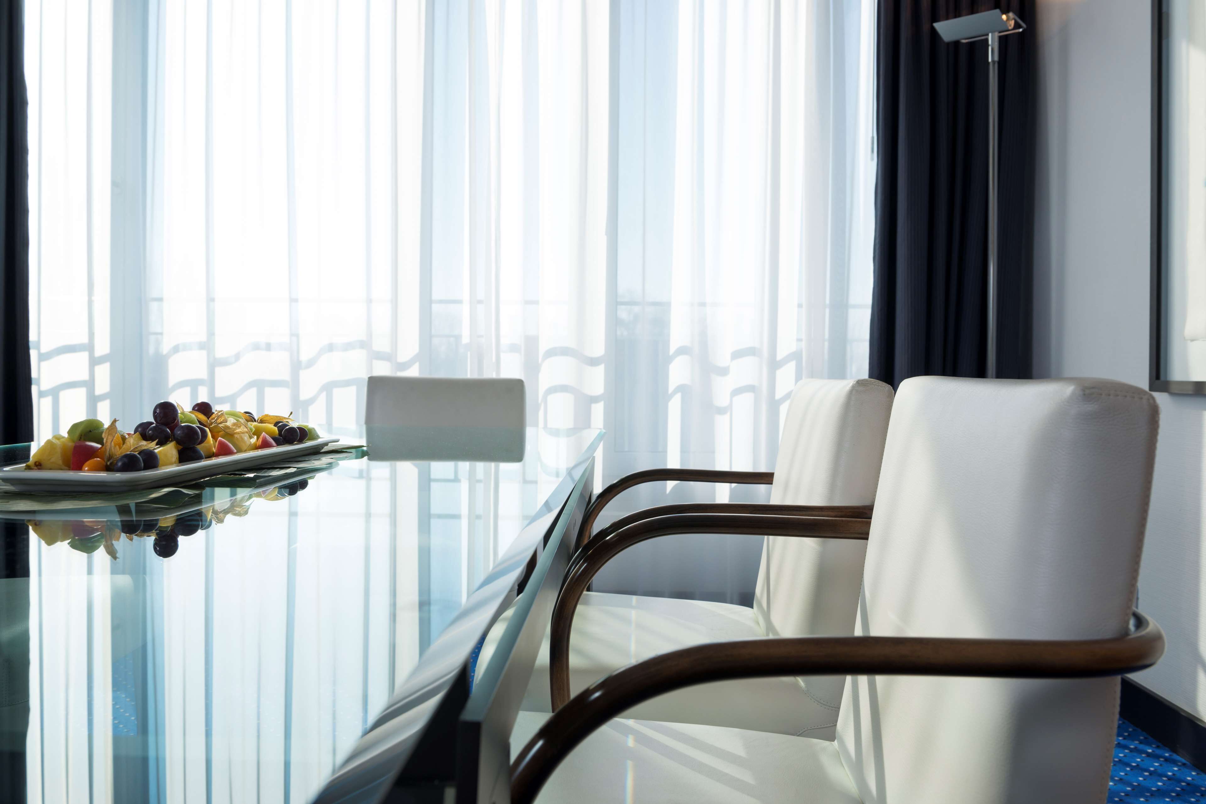 Una habitación de hotel con una mesita de vidrio, una bandeja de frutas y una silla blanca.