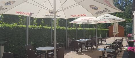 An outdoor patio area with tables and chairs under large umbrellas, some with visible brand names.