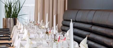 A long dining table set with white tablecloths, candles, and glassware, with a large window and curtains in the background.