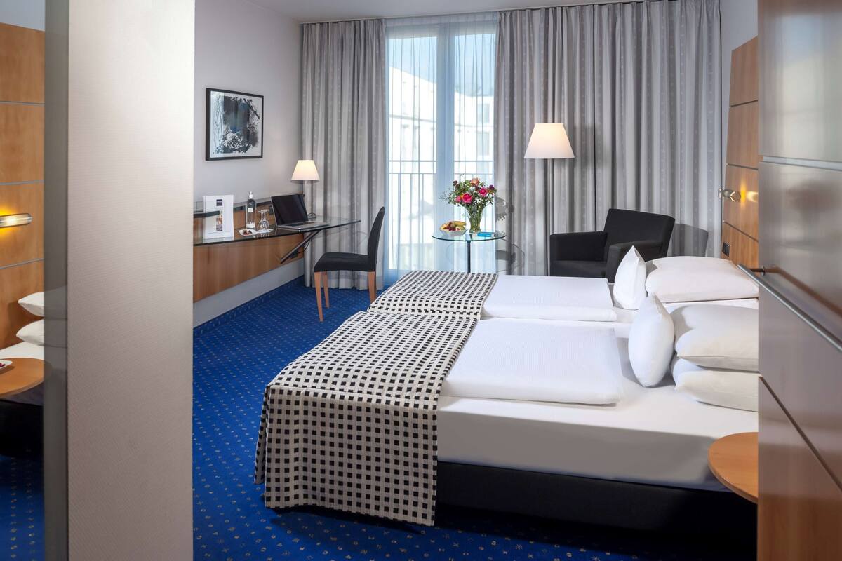 A hotel room with a bed, a desk, a chair, a small table with flowers, and a lamp.