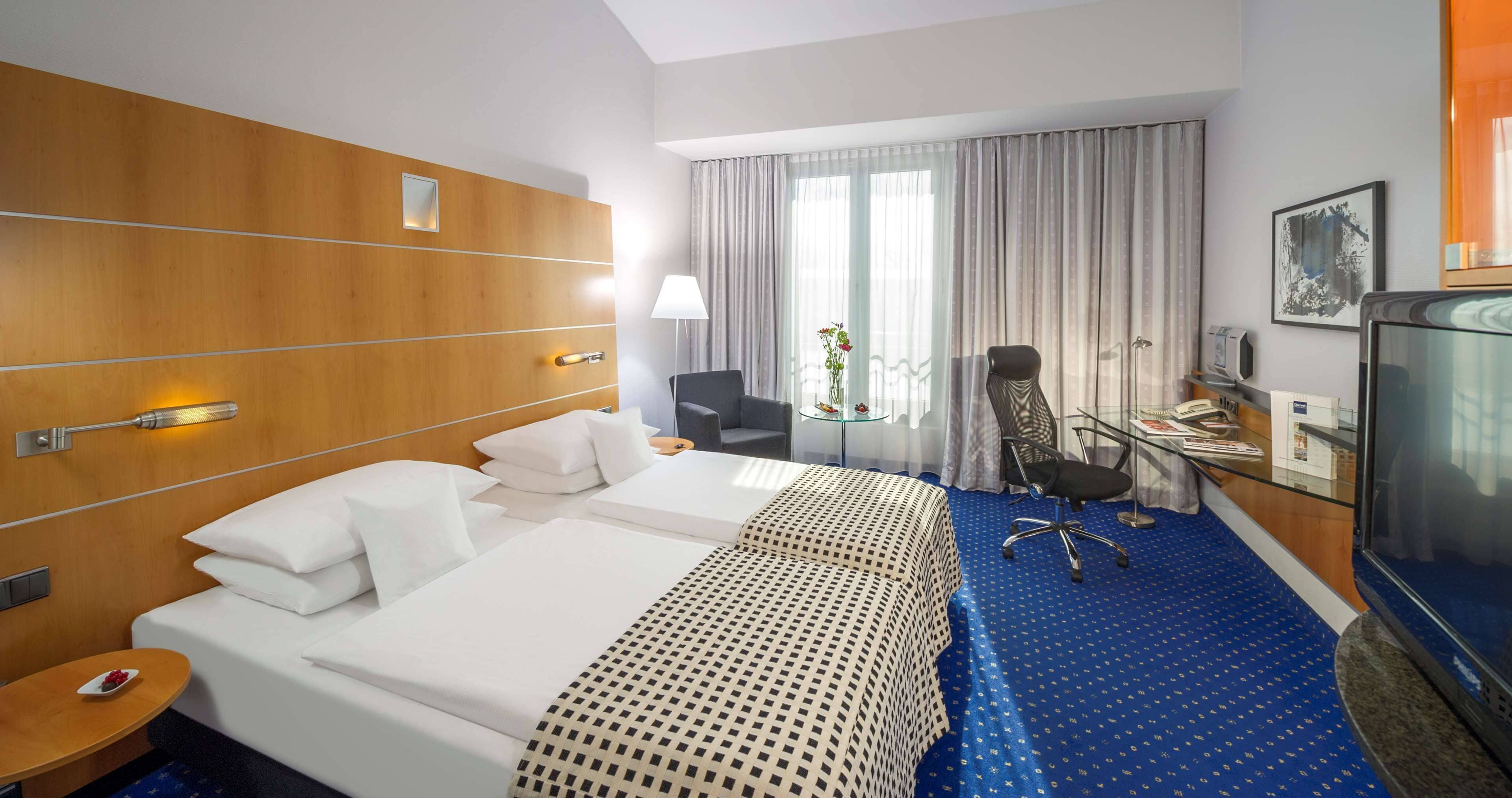 A hotel room with a large bed, a desk, a chair, a TV, and a window with curtains.