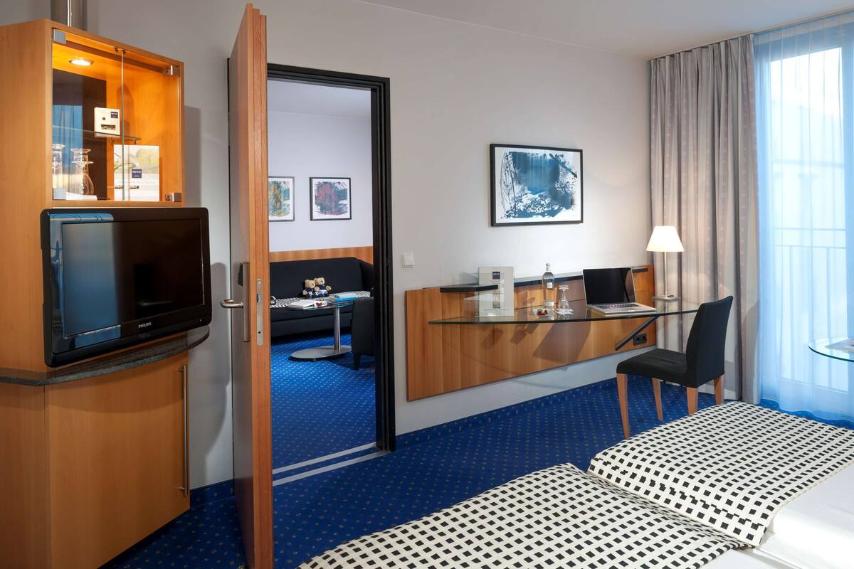 A hotel room with a bed, a desk with a computer, a TV, a chair, and a window with curtains.