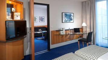 Quarto de hotel com cama, escrivaninha com computador, televisão, cadeira e janela com cortinas.