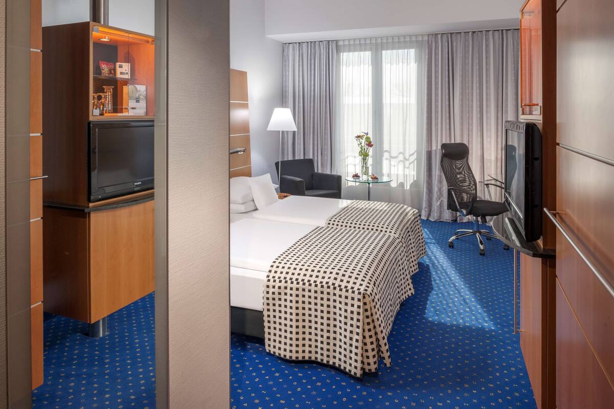 A hotel room with a bed, two chairs, a TV, a desk, and a minibar.