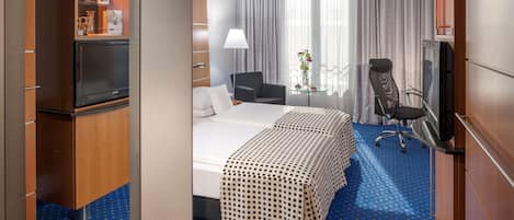 A hotel room with a bed, two chairs, a TV, a desk, and a minibar.