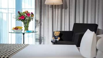 A hotel room with a bed, a chair, a lamp, a vase with flowers, and a bowl of fruit on a small table.
