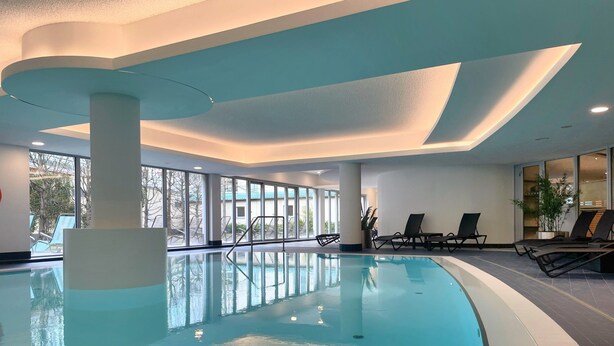 An indoor swimming pool with clear glass walls, lounge chairs, and a view of the outdoors. - Dorint Hotel Potsdam (Potsdam)