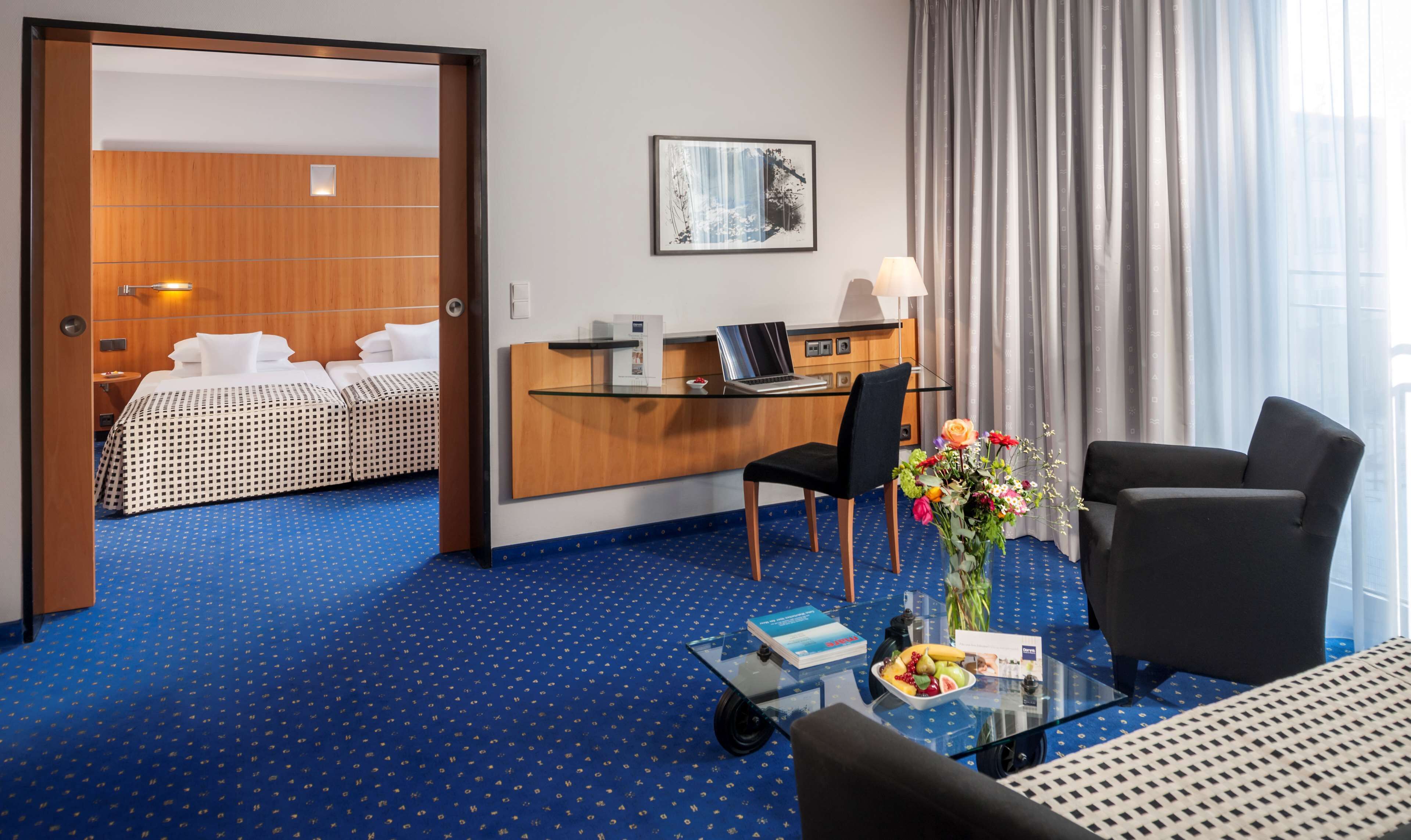 Une chambre d’hôtel avec un lit, un bureau, une chaise et une table basse en verre sur laquelle se trouvent un livre et des fruits.