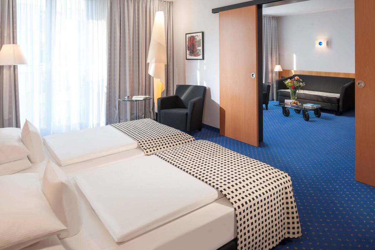 A hotel room with two beds, a sofa, a coffee table, and a large window with curtains.
