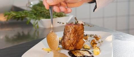 A chef's hand holding a spoon over a plate with a piece of meat, vegetables, and a sauce.