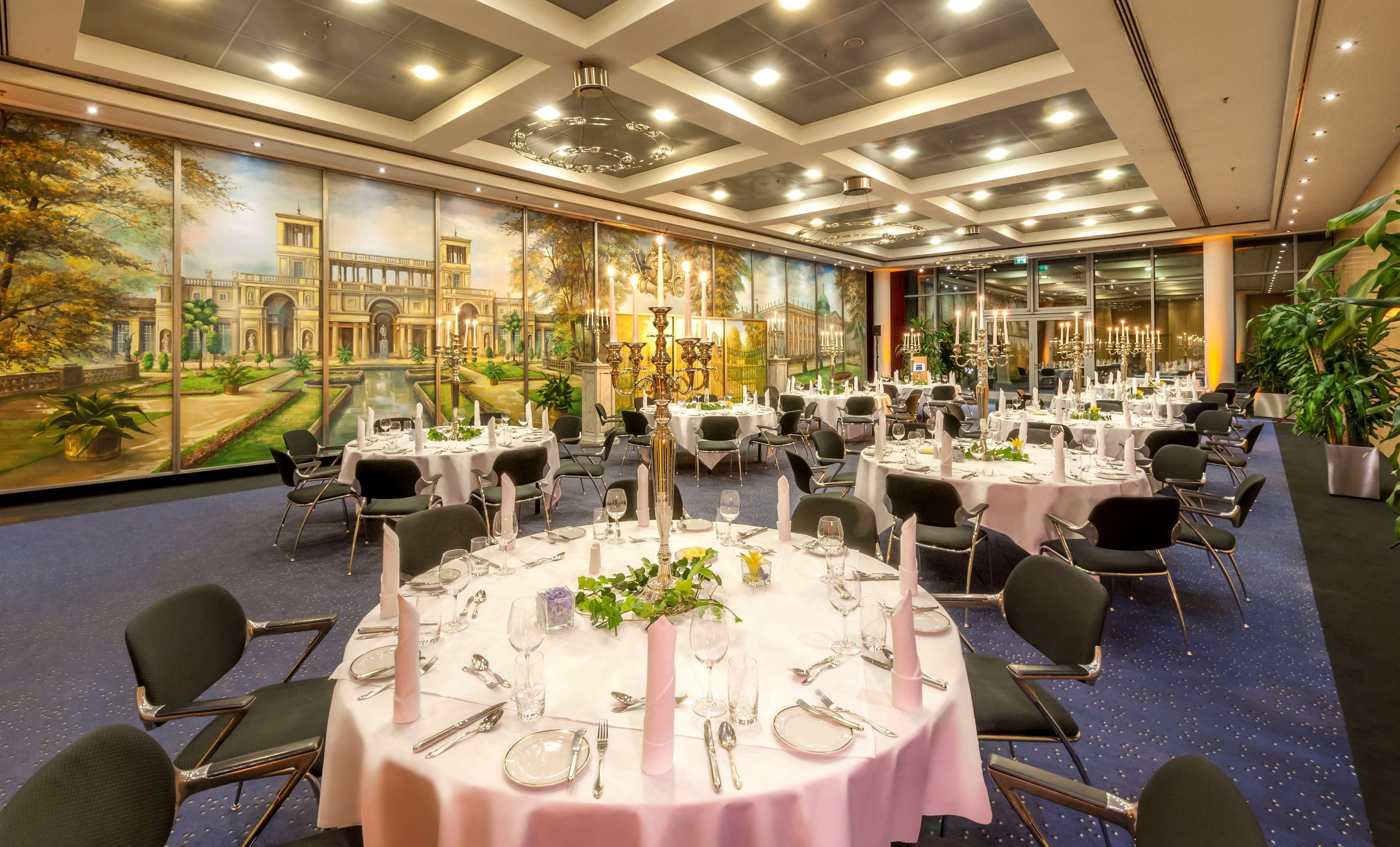 A banquet hall with round tables set for dining, a large mural on the wall, and a chandelier.