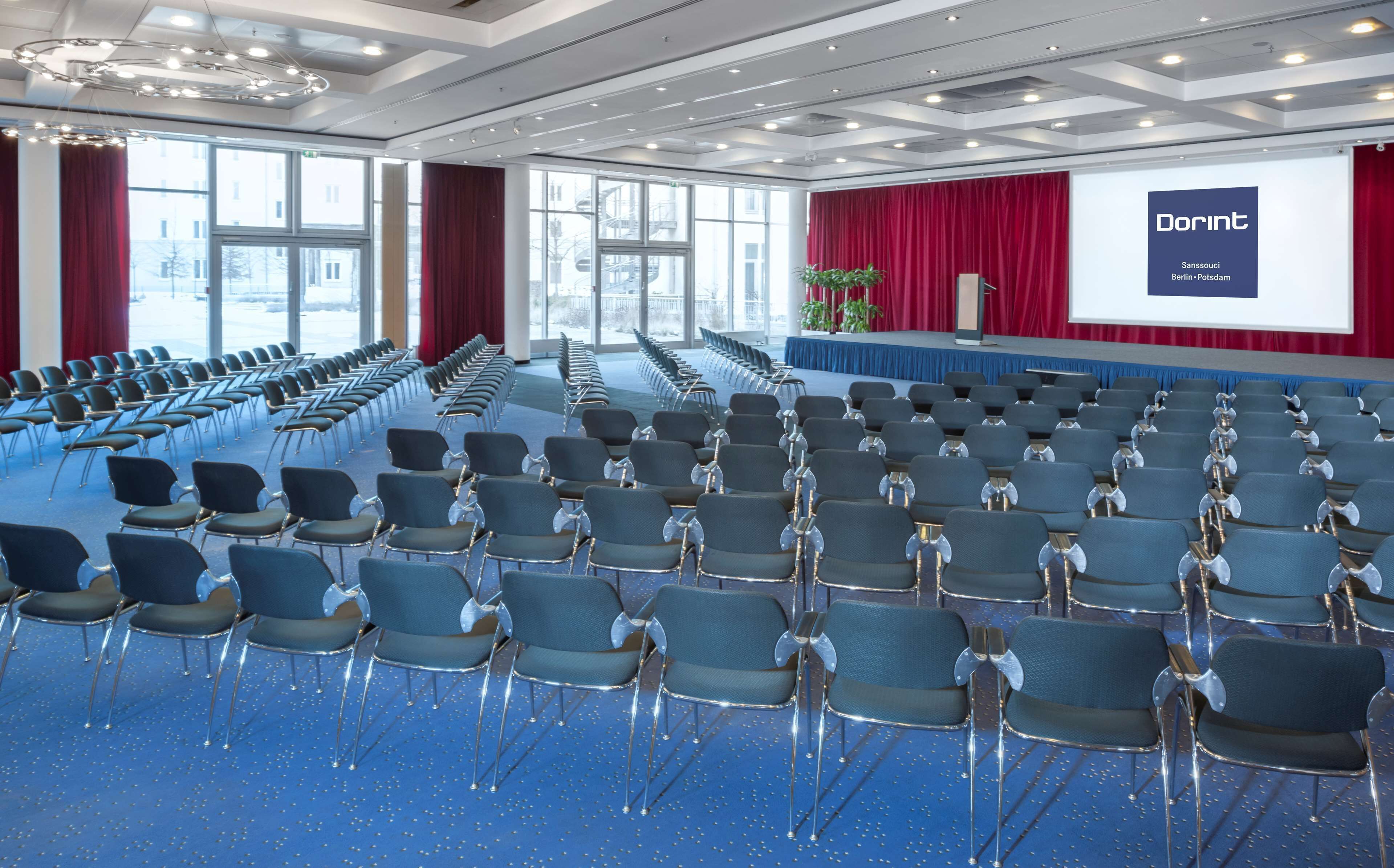 Un salón de conferencias con filas de sillas, un escenario con una pantalla y ventanas amplias.
