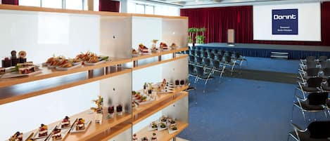 Un salón de conferencias con un escenario, filas de sillas y un bufé con diversos postres.