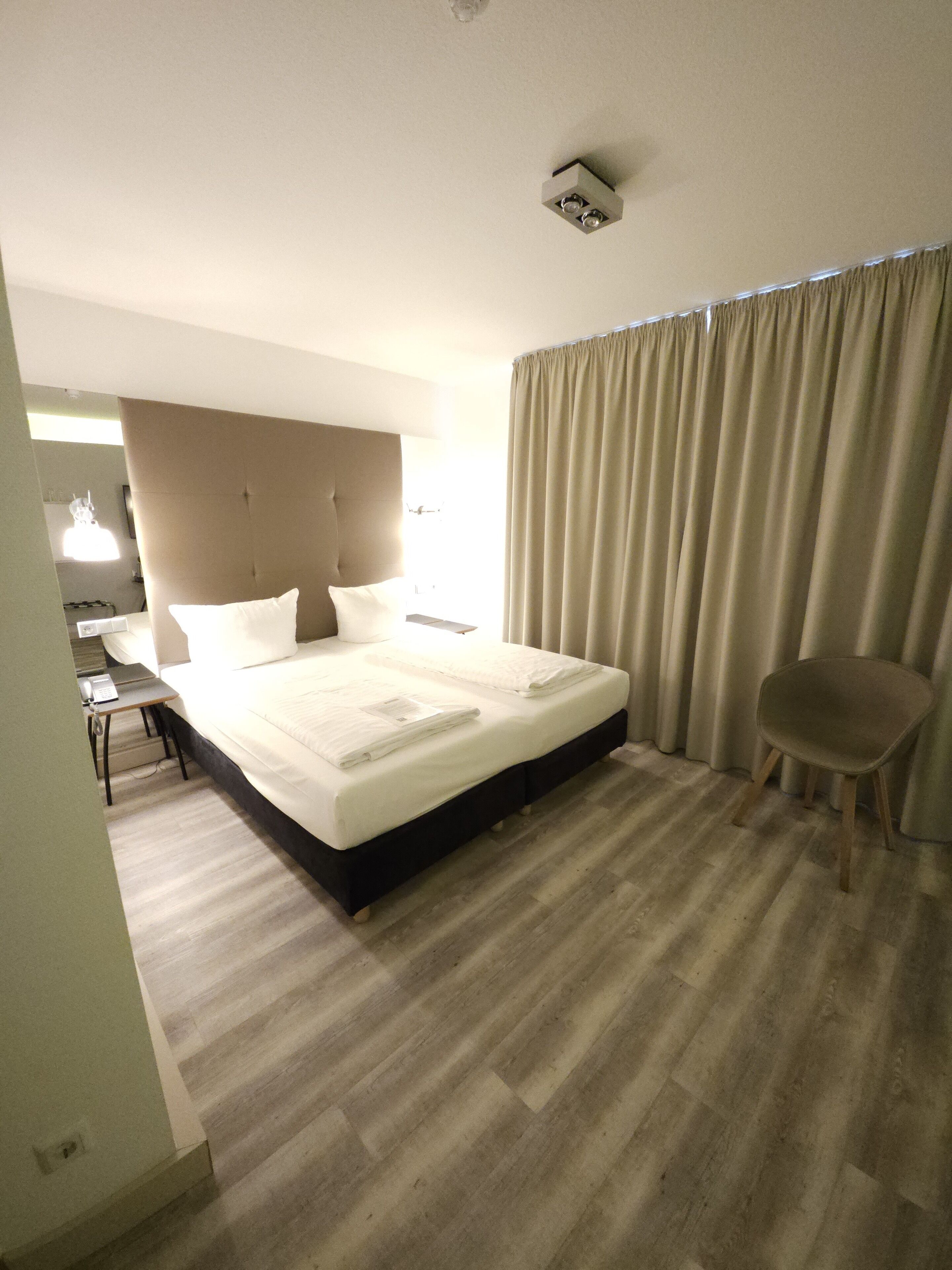 Quarto de hotel com uma cama grande, uma mesa de cabeceira com abajur, uma cadeira e luminária no teto.
