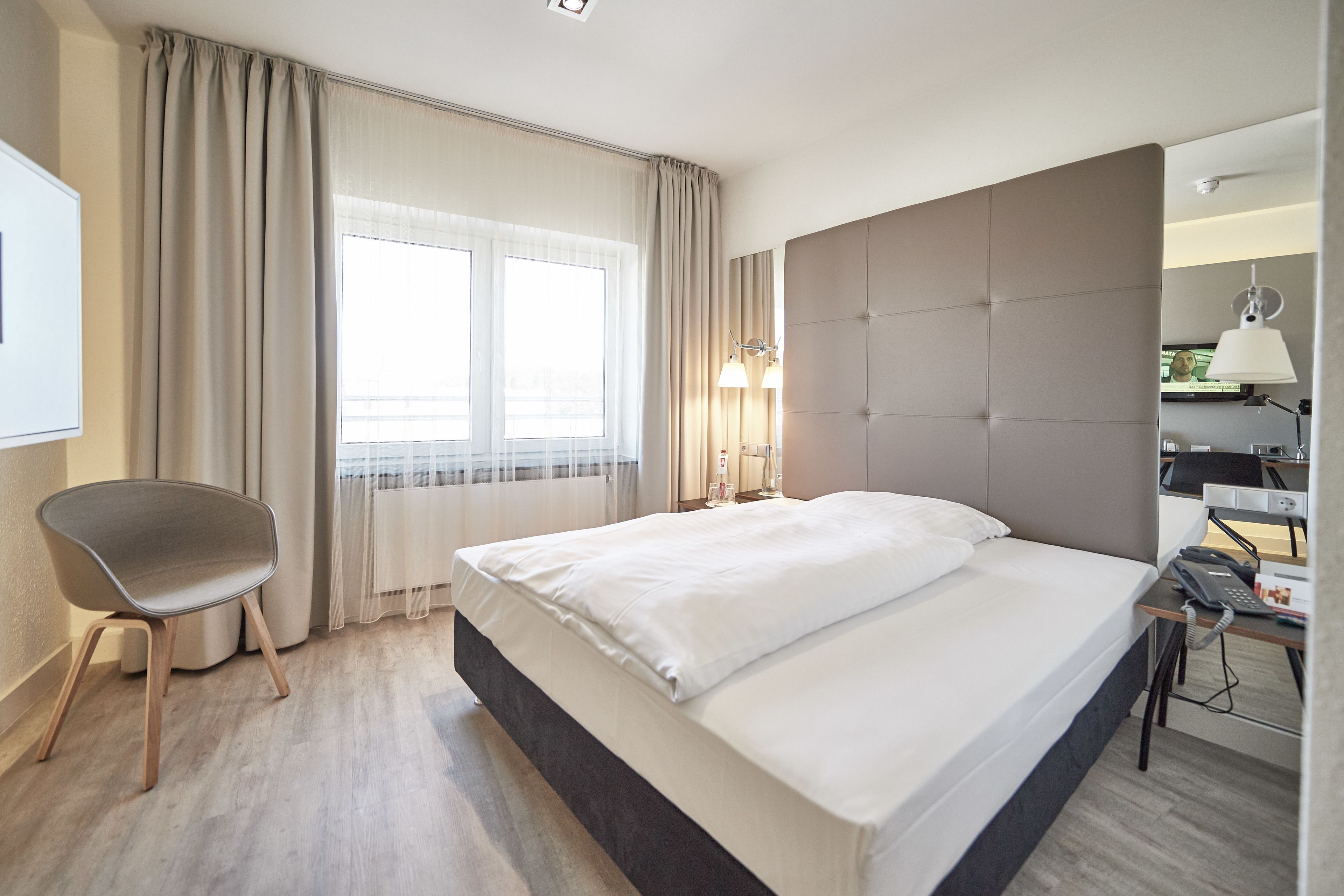 Hotellihuone, jossa on suuri sänky, työpöytä lampun kanssa, tuoli ja ikkuna, jossa on verhot.