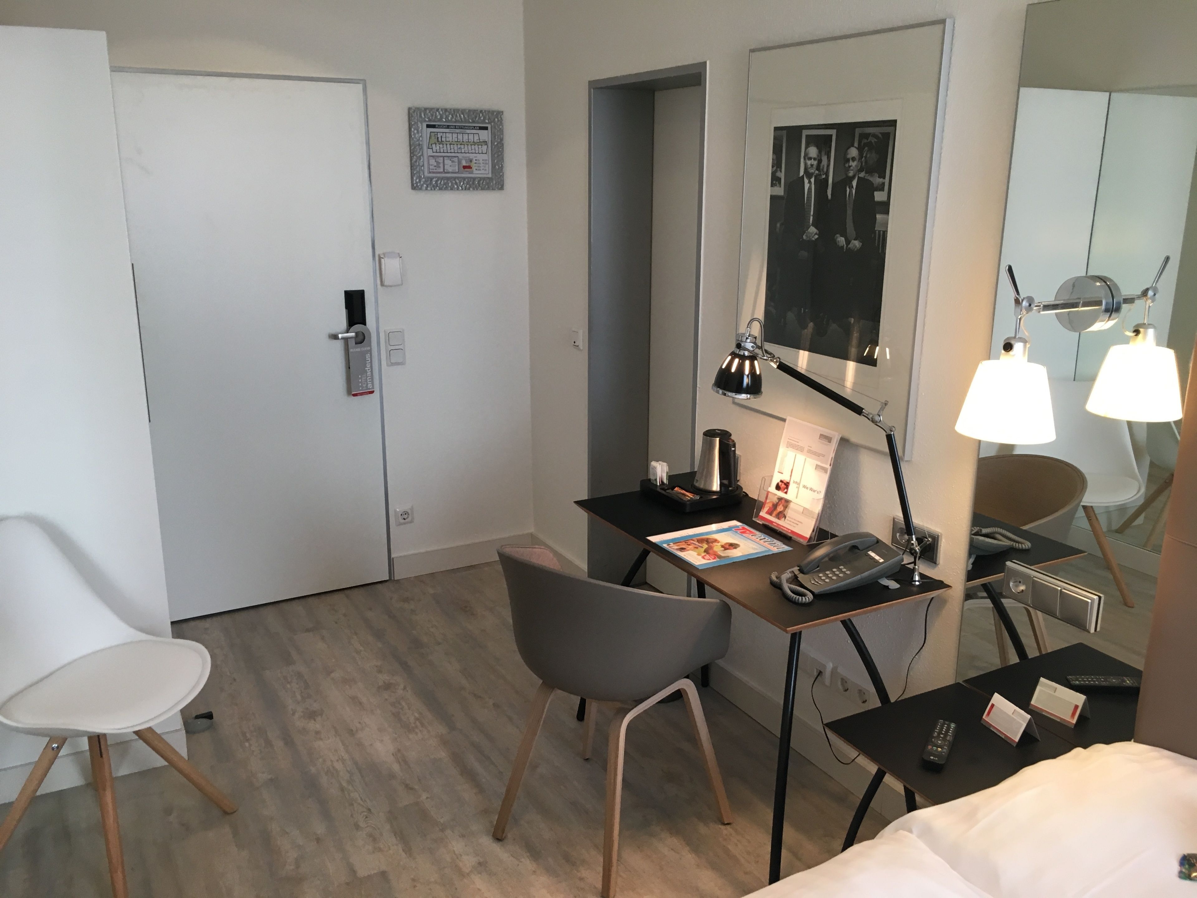 A hotel room with a desk, two chairs, a bed, a mirror, a lamp, a telephone, and a small table with a remote control.