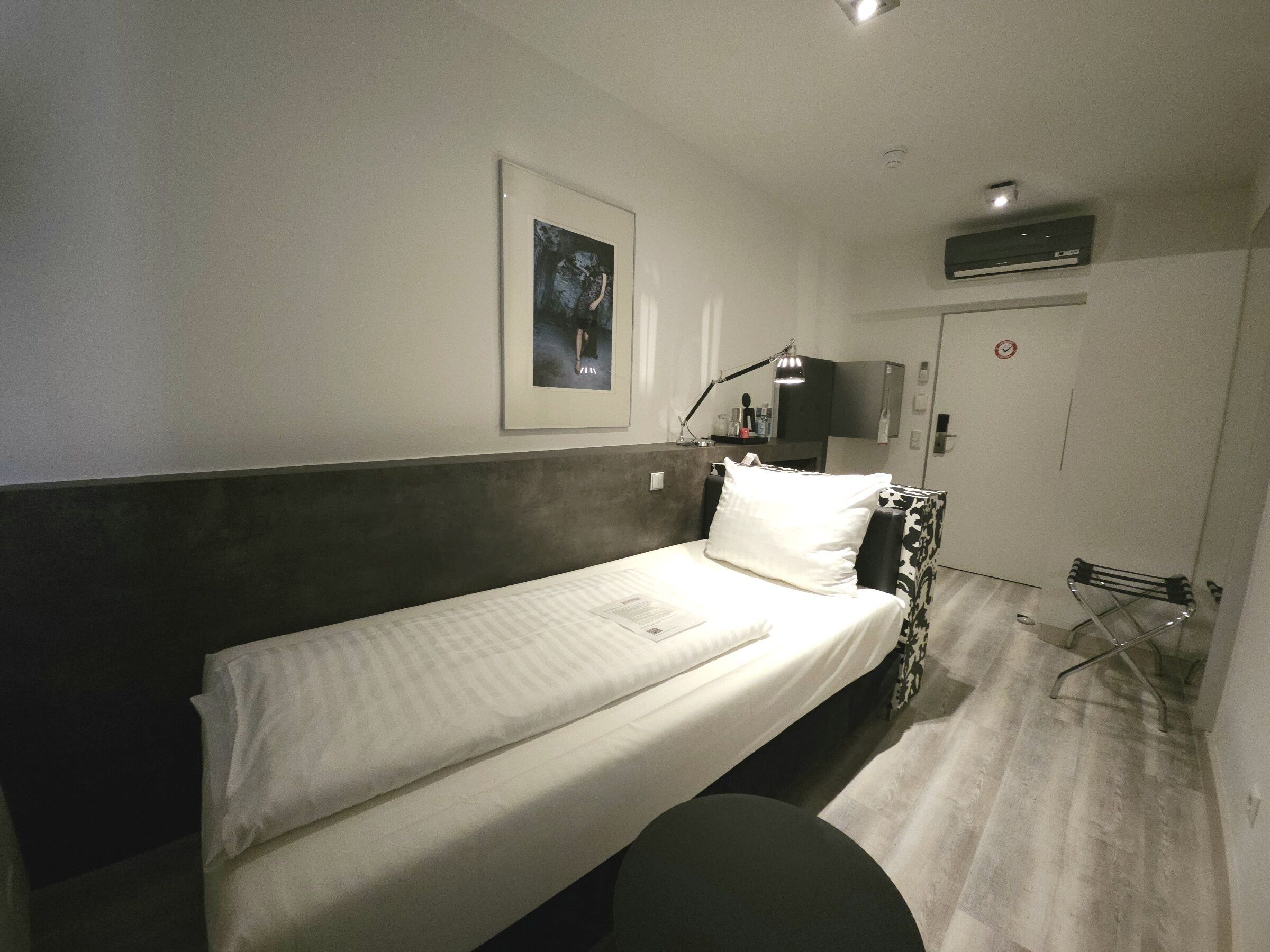 A single bed with a headboard, a bedside table with a lamp, a framed picture on the wall, and a door with a no-smoking sign.