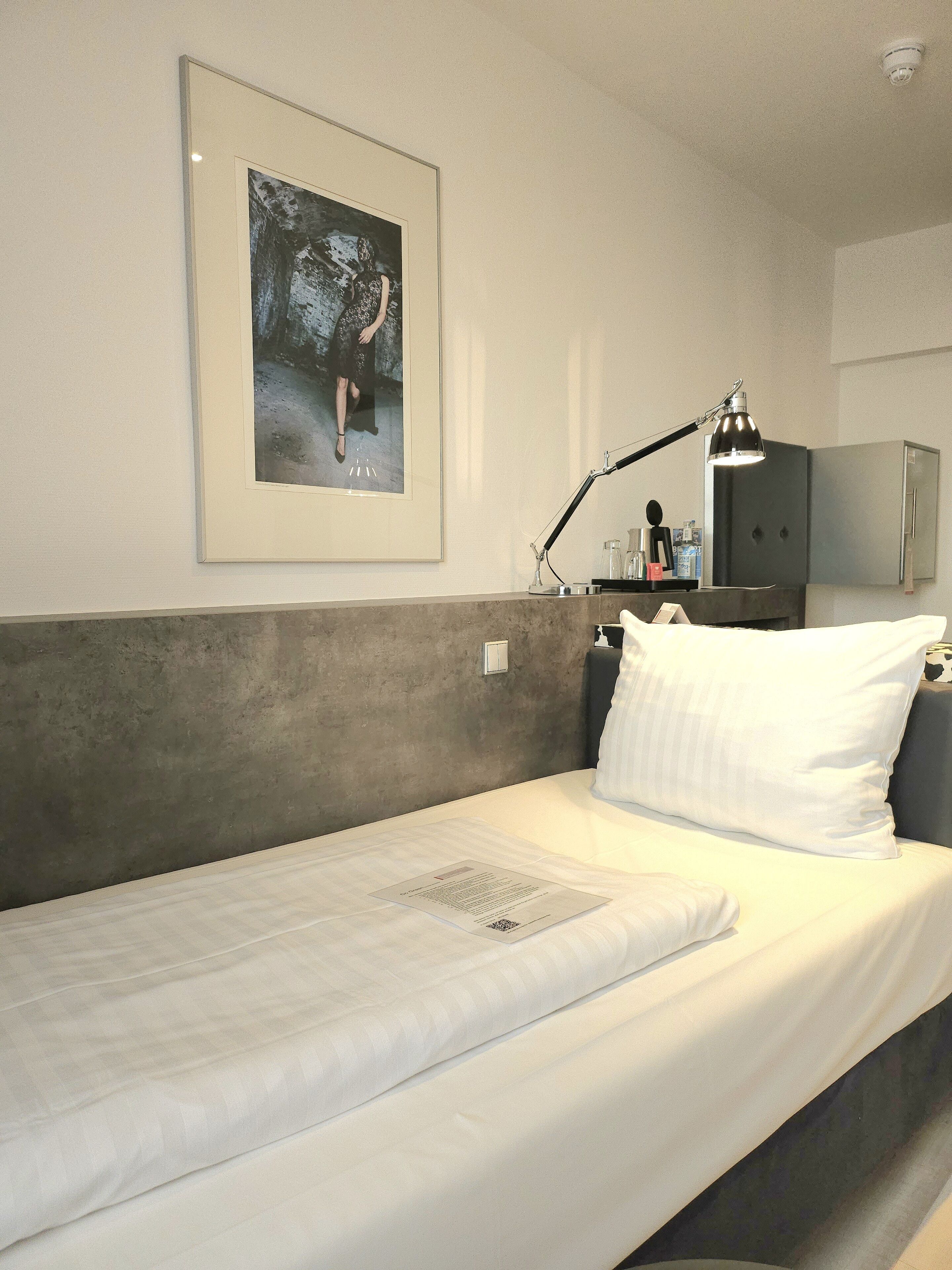 A single bed with white bedding, a framed picture on the wall, a desk with a lamp, and a wardrobe.