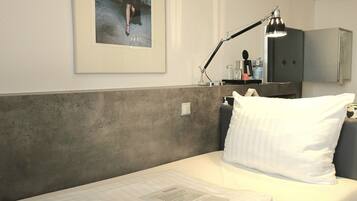 A single bed with white bedding, a framed picture on the wall, a desk with a lamp, and a wardrobe.