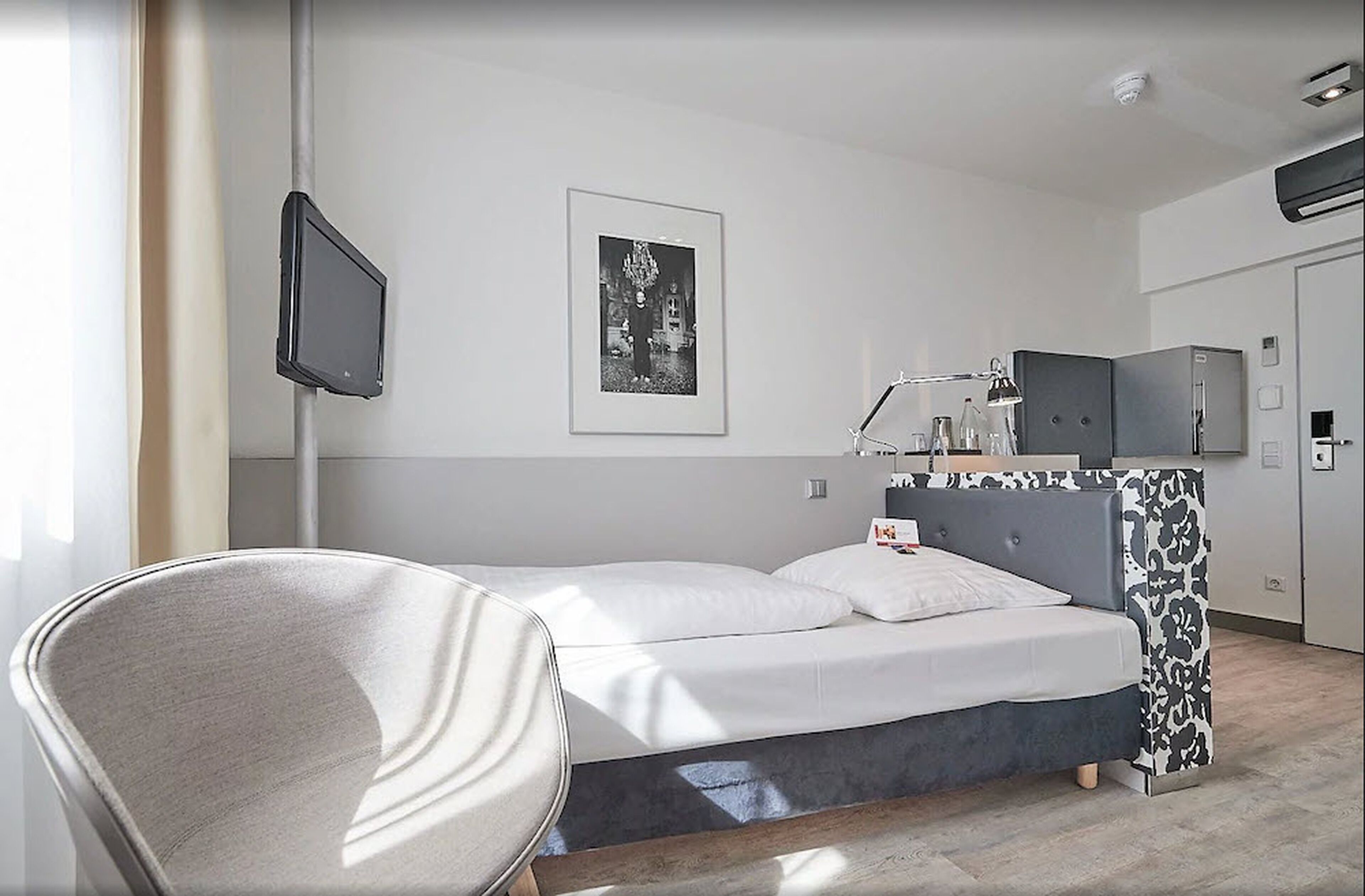 Een moderne hotelkamer met een bed, een stoel, een bureau met een lamp, een televisie en een ingelijst kunstwerk aan de muur.