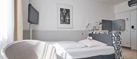 Moderni hotellihuone, jossa on sänky, tuoli, lamppuvarusteinen työpöytä, televisio ja seinällä kehystetty taulu.