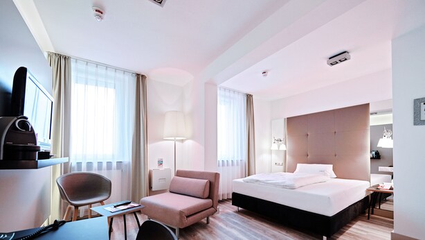 A hotel room with a bed, a desk, a chair, a lamp, and a television. - Hotel Amadeus (Hannover)