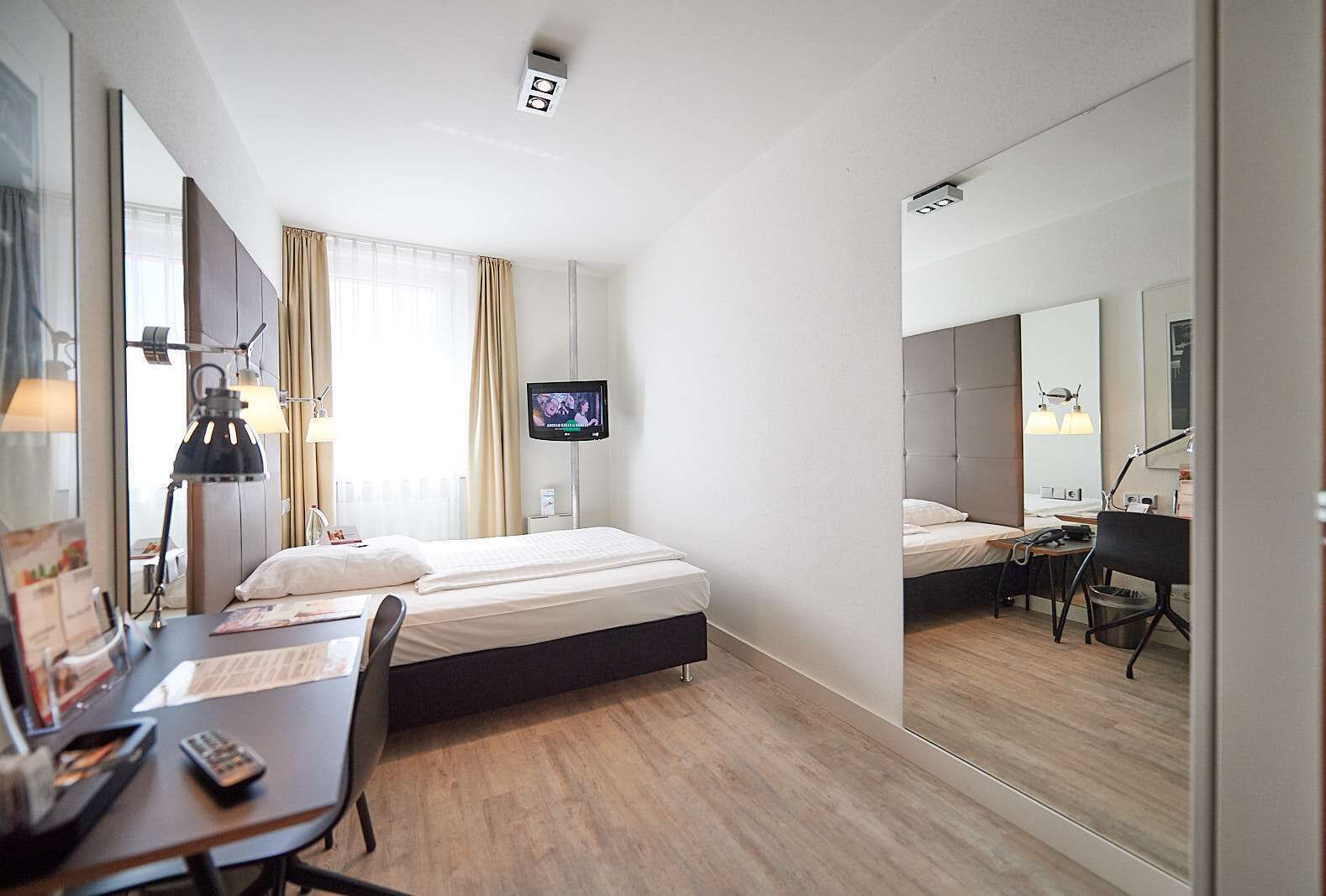 Quarto de hotel com cama, escrivaninha, cadeira, televisão e um espelho grande.
