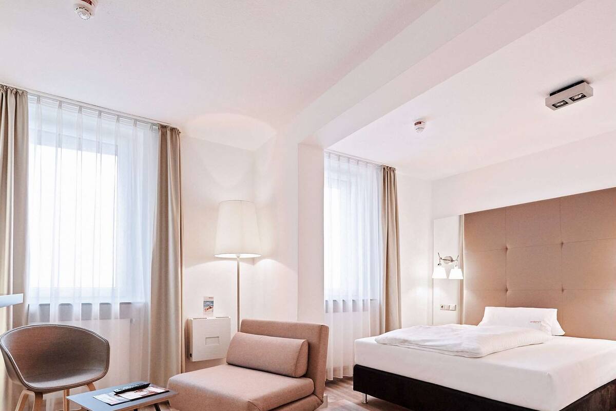 A hotel room with a bed, a chair, a desk, a lamp, and a window with curtains.
