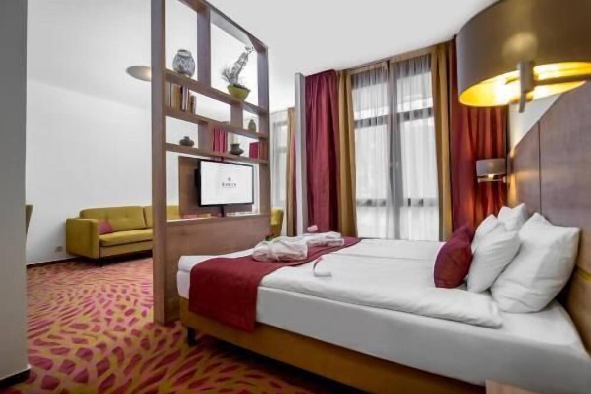 A hotel room with a bed, a television, a sofa, and a large window with curtains.