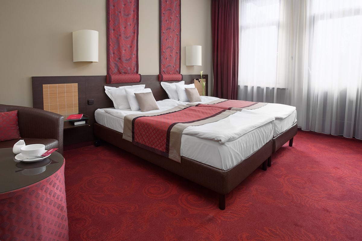 A hotel room with a large bed, a small table with a red tablecloth, a brown leather chair, and a red patterned carpet.