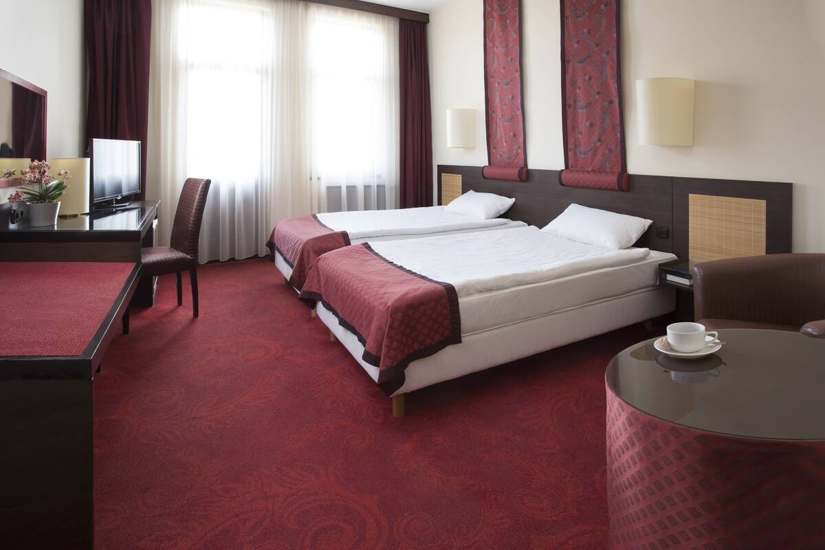 A hotel room with two beds, a desk with a TV, a chair, a round table with a cup, and a large window with curtains.