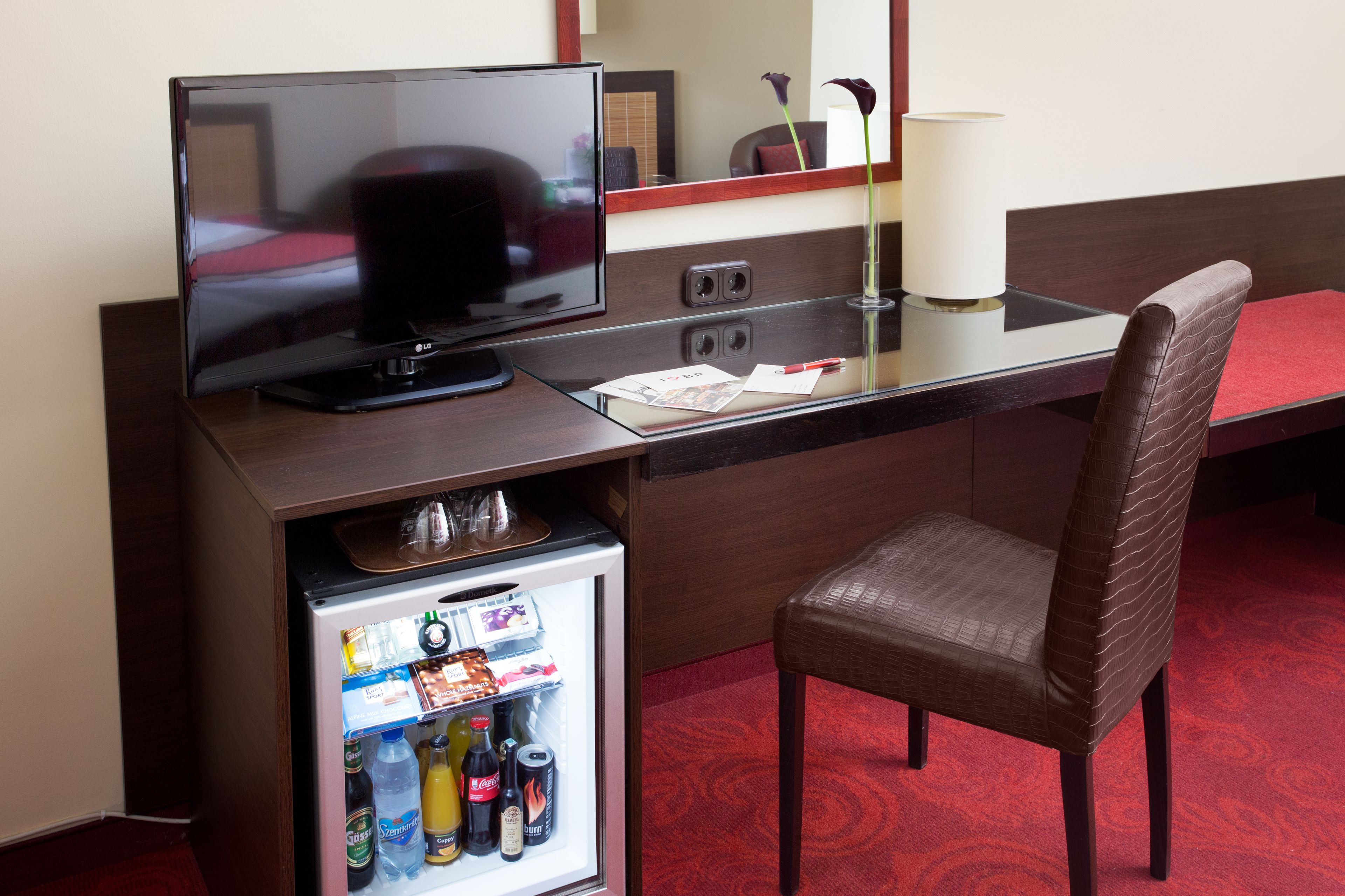 Habitación de hotel con televisión, minibar, escritorio, una silla y alfombra roja.