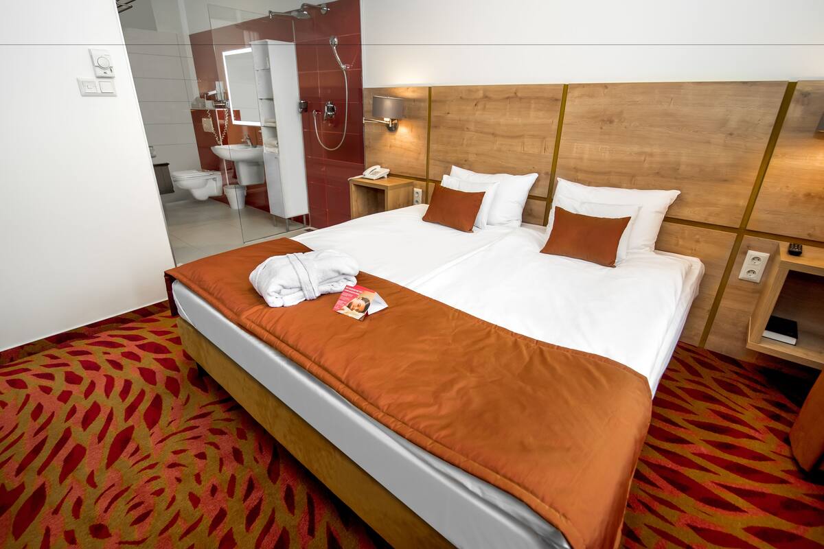 A hotel room with a large bed, a wooden headboard, a bathroom visible through a glass partition, and a patterned carpet.