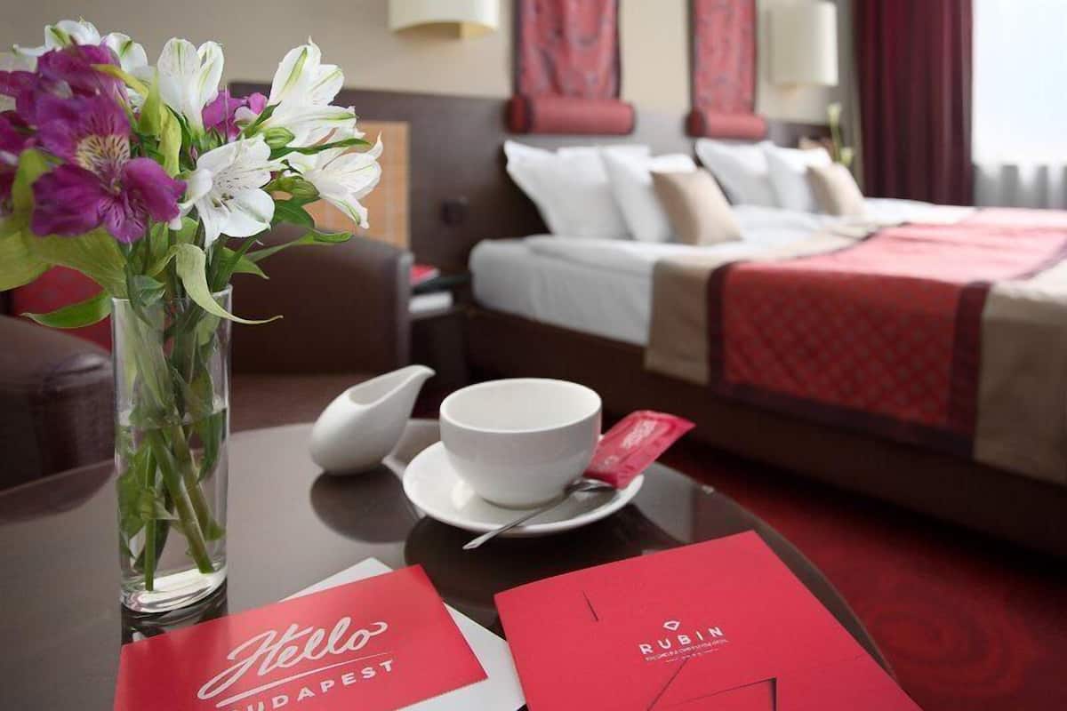 Habitación de hotel con cama, un jarrón de flores, taza y platillo, y dos tarjetas rojas.