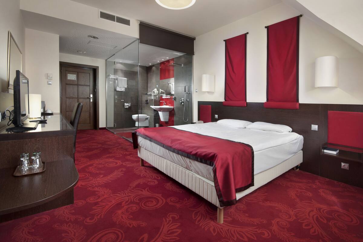 A hotel room with a bed, a desk, a mirror, a shower area, and a red carpet.