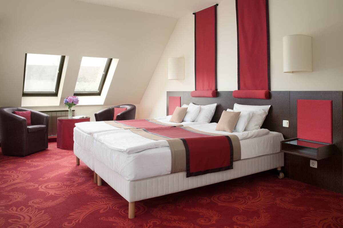 A hotel room with a large bed, two armchairs, a small table with a vase of flowers, and a red carpet.