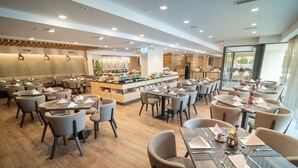 Un comedor moderno con varias mesas y sillas, una estación de buffet central y vistas al exterior a través de amplios ventanales.