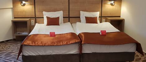 A double room with two beds, each with a brown headboard and white bedding. There are two bedside tables with lamps, and a patterned carpet on the floor.